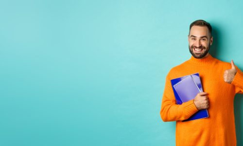 Education. Smiling bearded man holding notebooks and smiling, going on courses, showing thumbs-up in approval, like something, standing over turquoise background.